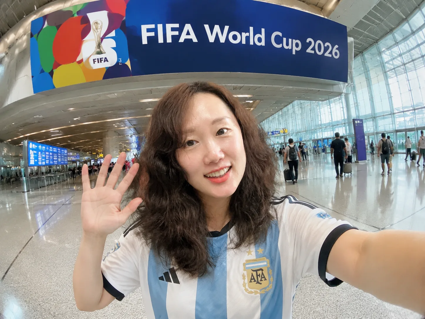 A smiling young woman in an Argentina striped jersey waves for a selfie at a busy airport.